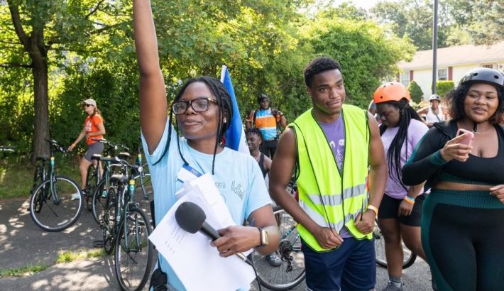 Three young black people standing in front of a row of parked bicycles. The woman in the center is wearing a blue t-shirt and glasses and holding a microphone in one hand and raising her other hand high. 