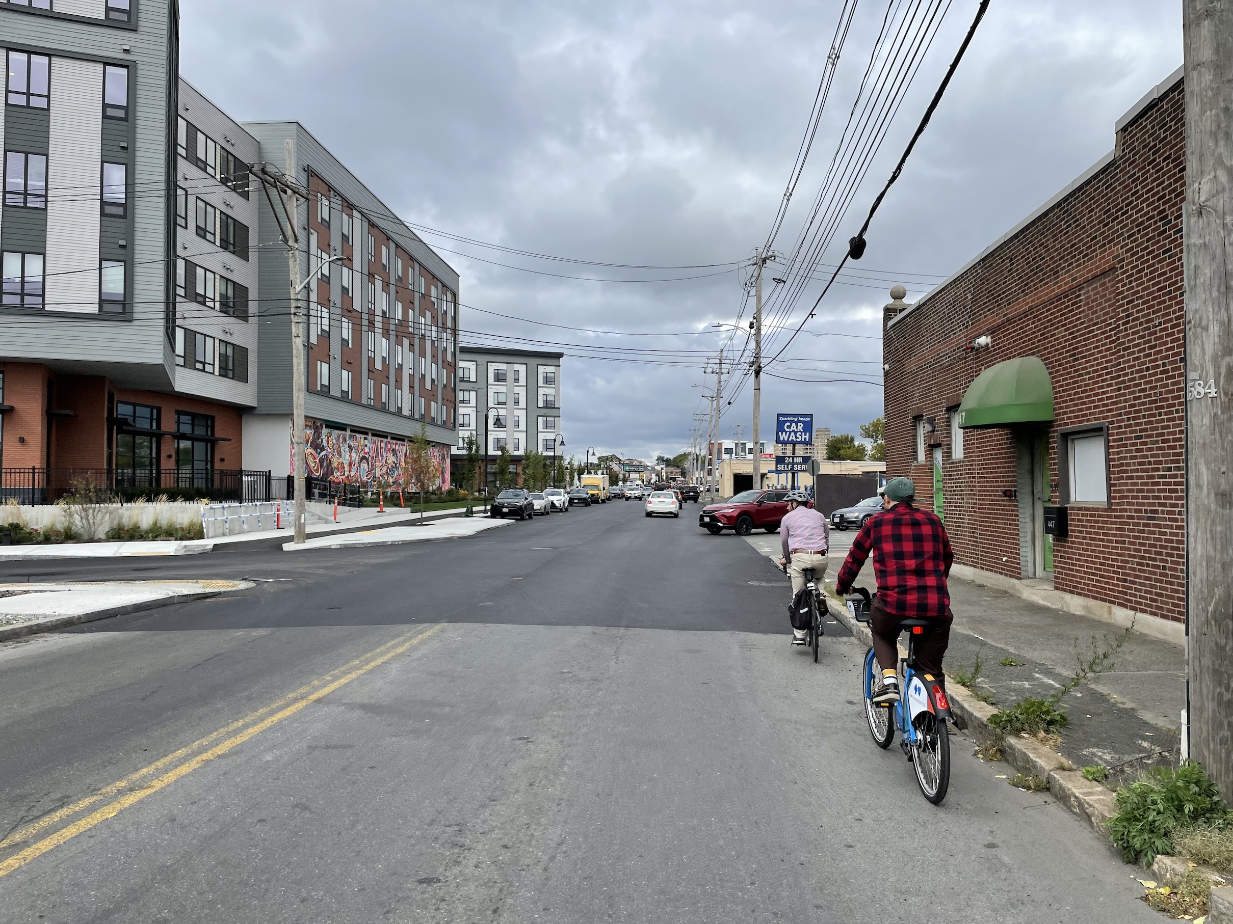 Two people on bikes ride along a city street next to a new 5-story apartment building along the left side of the street. The right side of the street has an old single-story brick warehouse building and power lines above. 