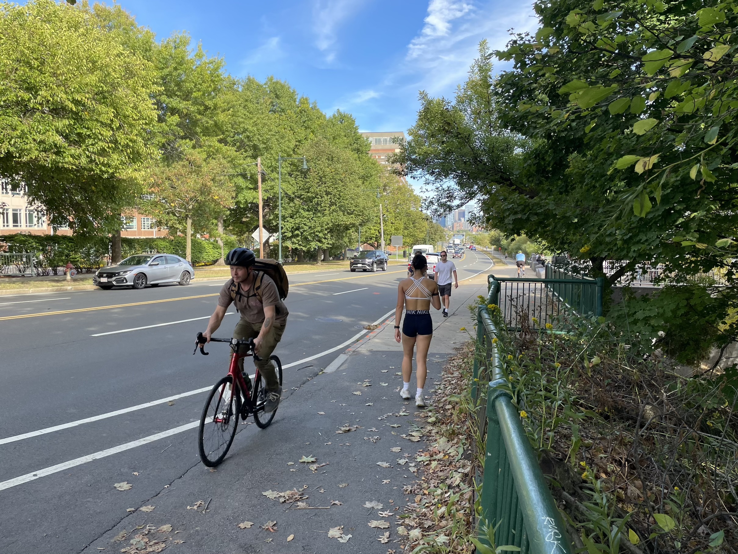 Several pedestrians and a person on a bike pass each other on a narrow sidewalk next to a wide multi-lane highway. 