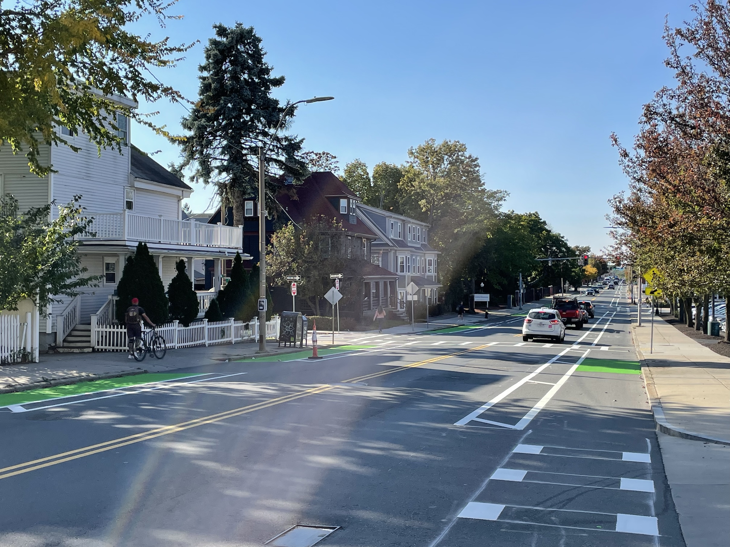 A city street under a sunny blue sky. The street features two bike lanes along each curb on either side of the street with hatched lines to buffer the bike lane from moving car traffic in the middle of the street.