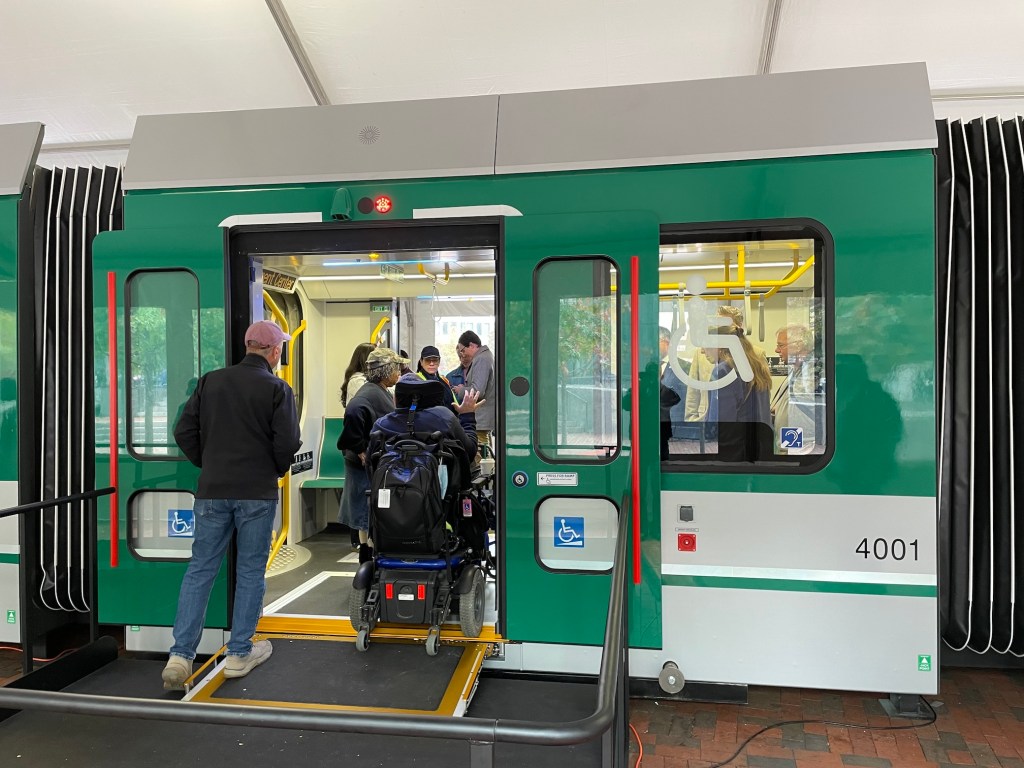 A person in a wheelchair uses a small ramp to cross the threshold of a MBTA Green Line train next to a window with a large wheelchair icon.