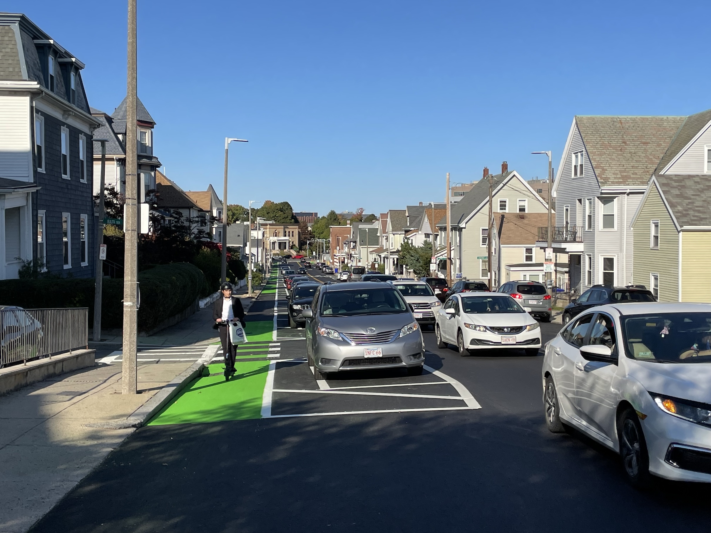 A recently-paved street lined with 2 and 3-story houses with gable roofs. The street features a bike lane on the left, with a man riding a scooter towards the camera. Between the bike lane and moving traffic in the center of the street is a row of parked cars.