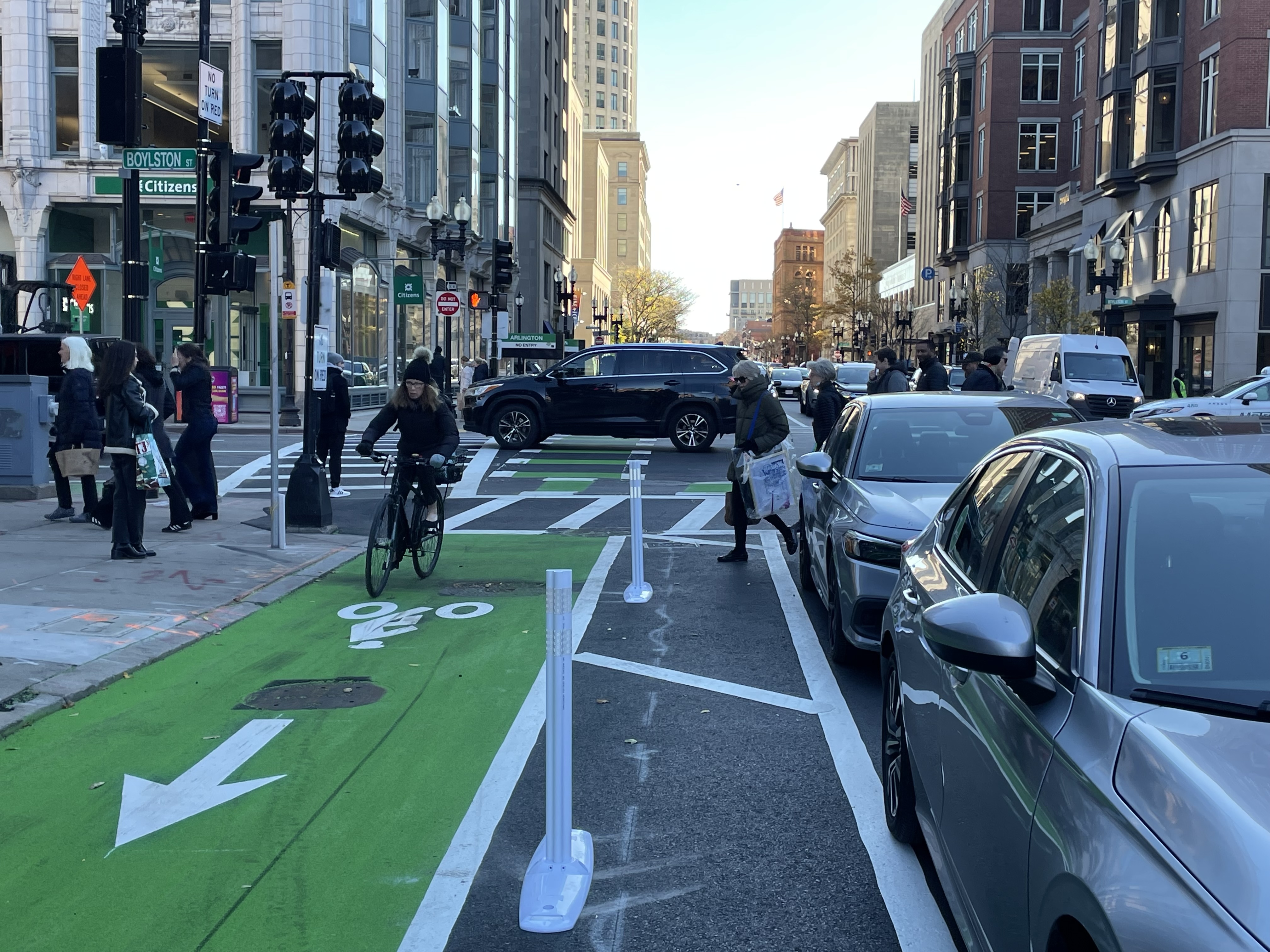 A person rides a bike on a freshly-painted green bike lane next to some flexible-post bollards on a busy multi-lane city street lined with tall buildings