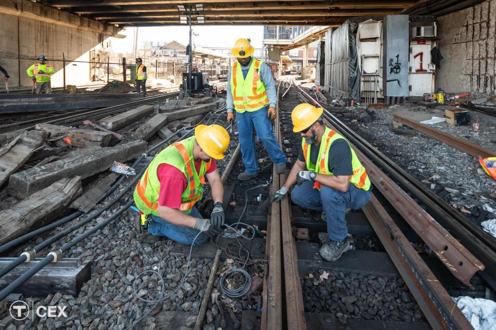 Three workers in helmets and fluorescent vests work on railroad tracks under a viaduct.