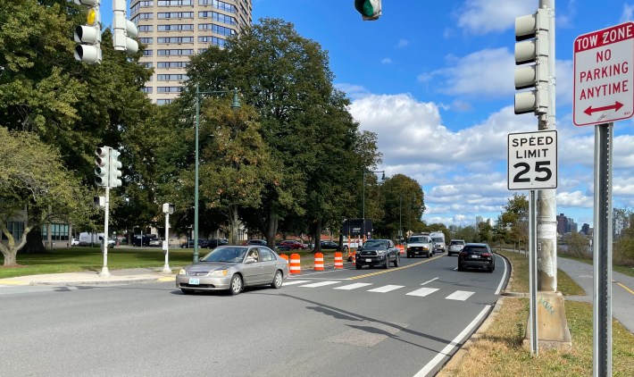 Cars drive along a curving multi-lane roadway lined with leafy trees. A high-rise apartment building is visible through the trees in the background; on the right side of the road in the foreground are a traffic light, a 25 mph speed limit sign, and a no parking sign.