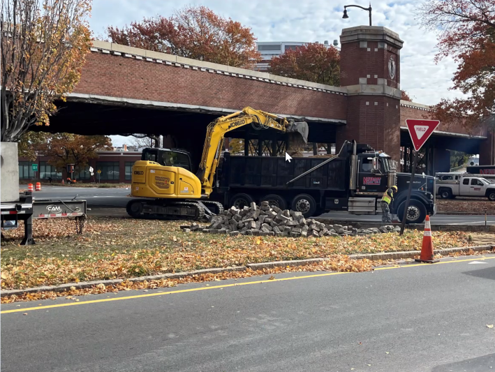 An excavator lifting rocks into a dump truck, demoing the sidewalk under the Reed Overpass.