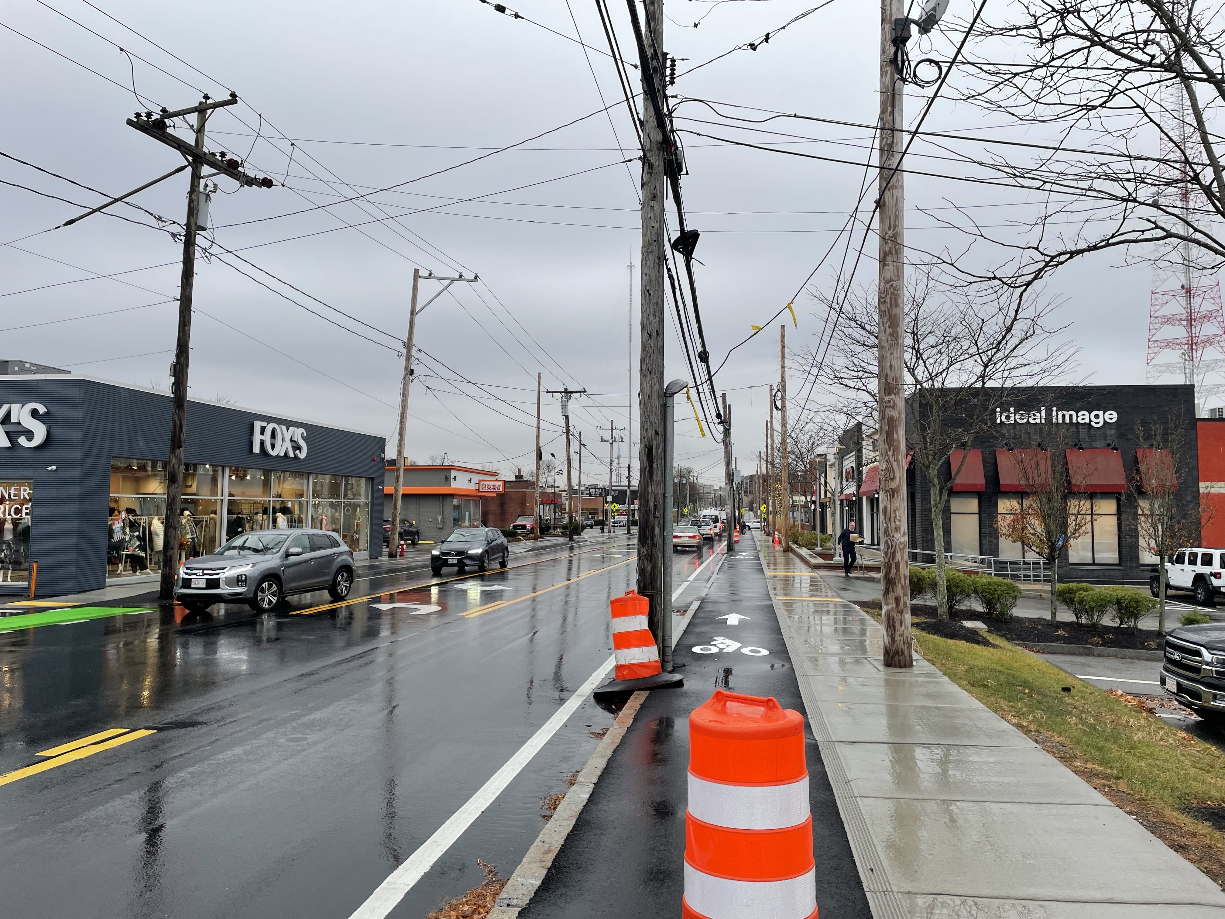 Dozens of wires suspended over a street lined with small businesses and orange construction barrels. The photo shows four rows of utility poles along the street. In the center, pairs of poles are located on the outside of the sidewalk and in the curb between the street's bike lane and roadway; on the left edge of the photo on the other side of the street are more poles.