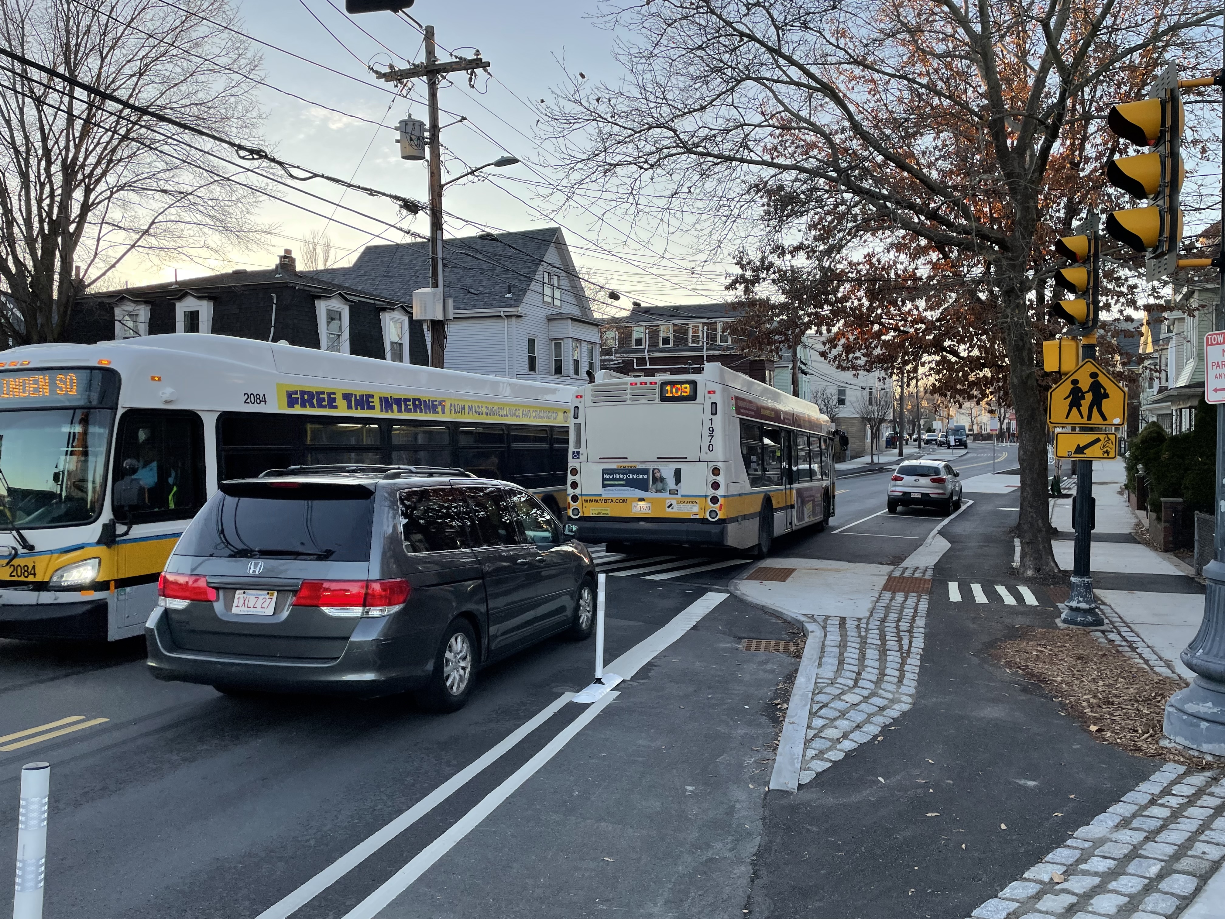 Two MBTA route 109 buses pass each other next to a traffic light on a two-lane street with sidewalk-level protected bike lanes