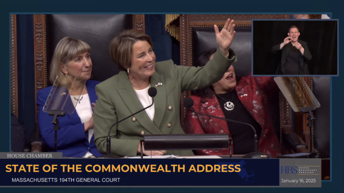 A woman with short brown hair in a green suit stands at a podium, smiling and waving, with two seated colleagues in colorful blazers behind her. Behind them is an ornate chair and a microphone stand, with an inset of an ASL interpreter on the screen.
