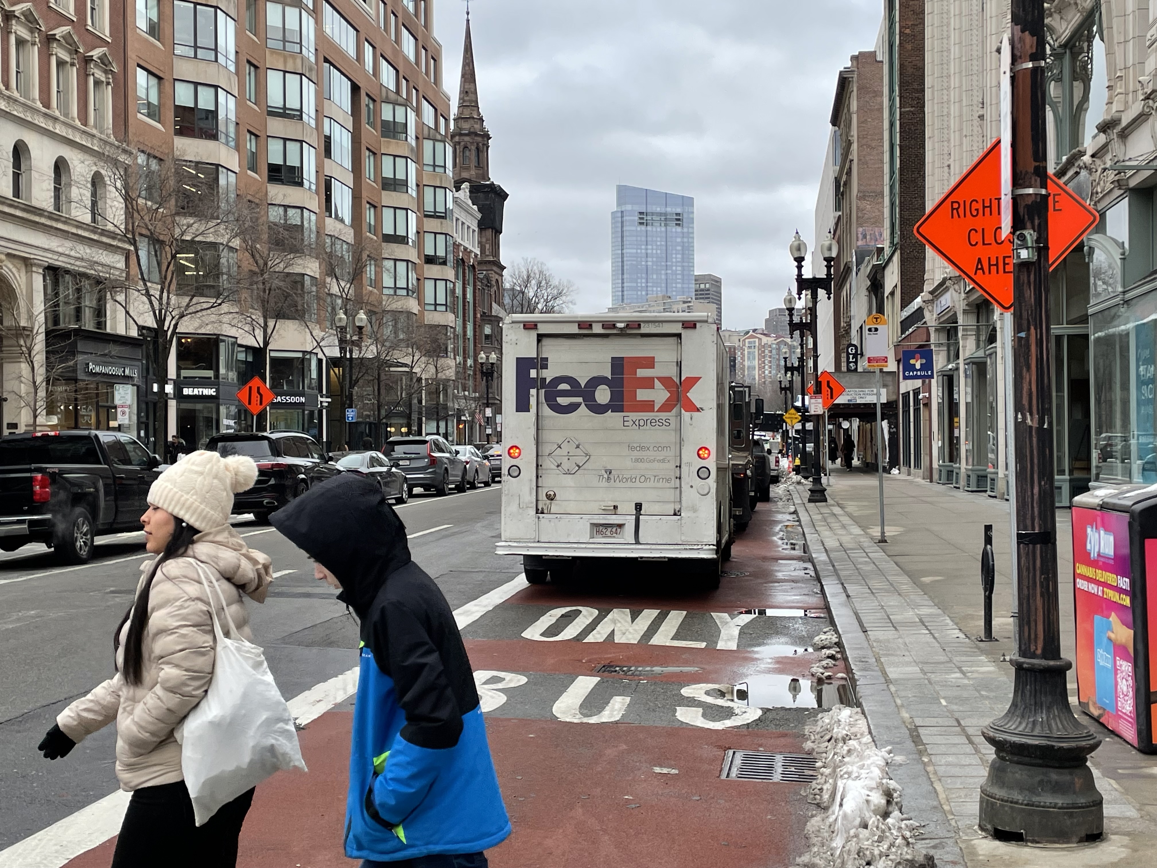 A FedEx truck and other illegally parked vehicles parked in the middle of a red bus lane marked with the words "ONLY BUS" on a city street lined with tall buildings. In the foreground, two people in winter coats cross the street.
