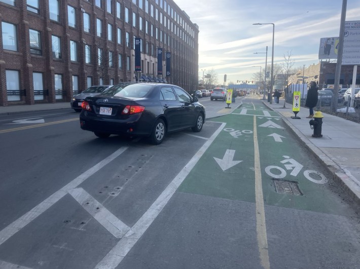 A wide city street next to a multi-story brick building at left. In the center of the photo is a two-way bike path divided from the adjacent car lanes with a pair of striped white lines.