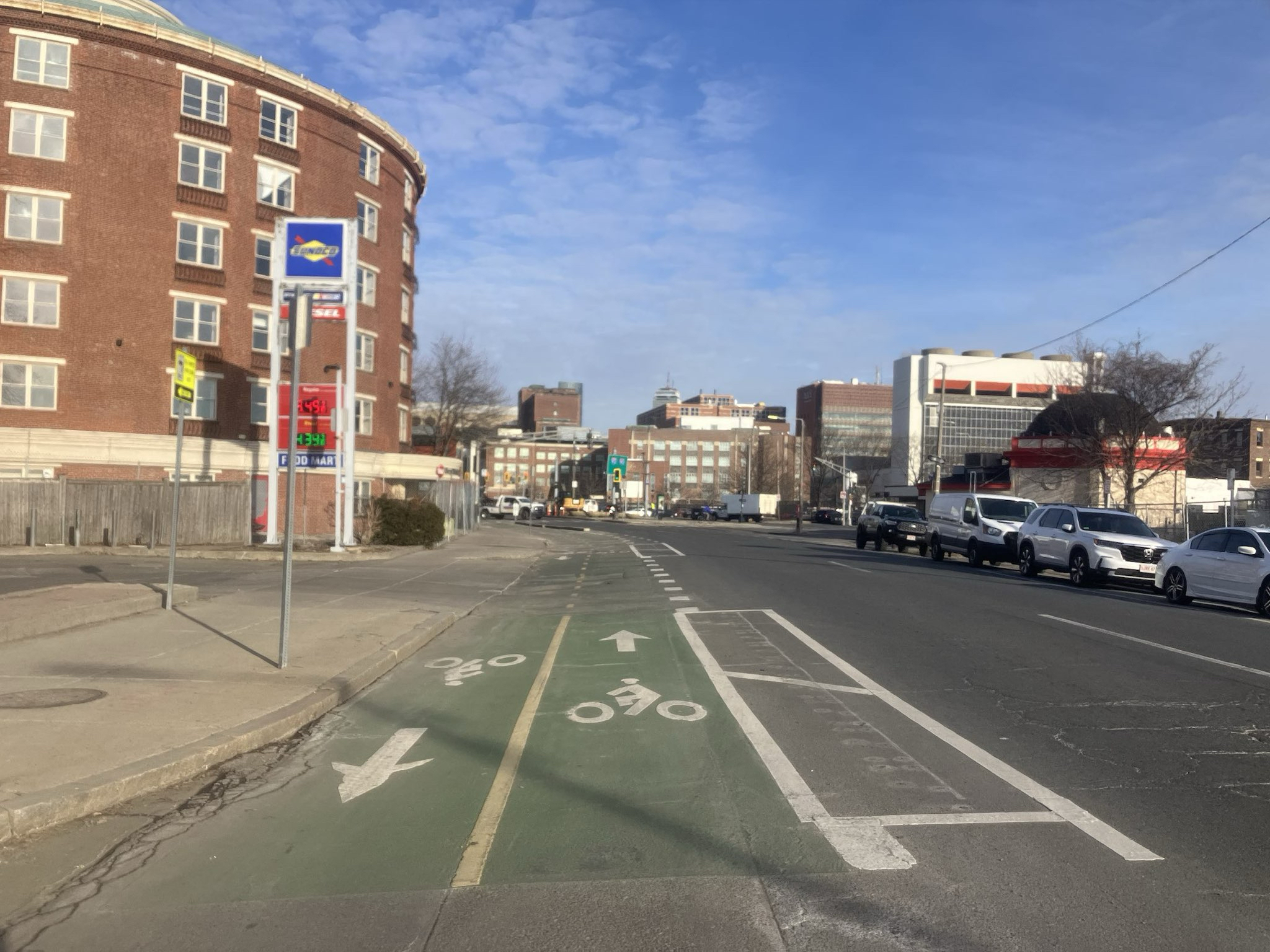 A wide city street next to a gas station and a cylindrical multi-story brick building. In the center of the photo is a two-way bike path divided from the adjacent car lanes with a pair of striped white lines.