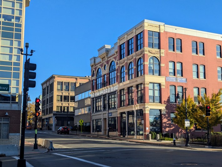 A photo of downtown Brockton, MA, showing a street corner with brick mid-rise buildings under a bright blue sky. A red-brick historic building labeled “Enterprise Building” stands out prominently.