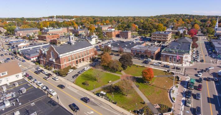 Aerial view of Needham Center, MA, with a large park, red-brick town hall, surrounding low-rise buildings, and parking lots. Tree-lined streets and suburban homes stretch into the distance.