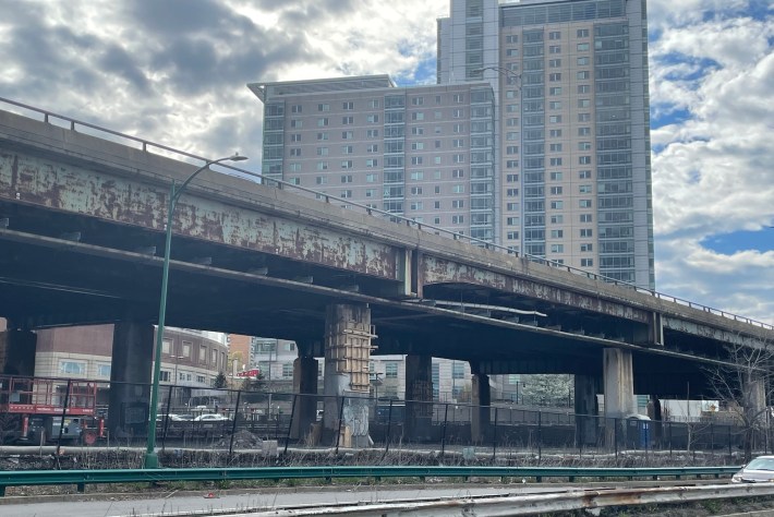 An aging highway viaduct, seen from below, with severely rusted green-painted steel beams that sit on tall rust-stained concrete columns with considerable damage. In the background is a high-rise apartment building.