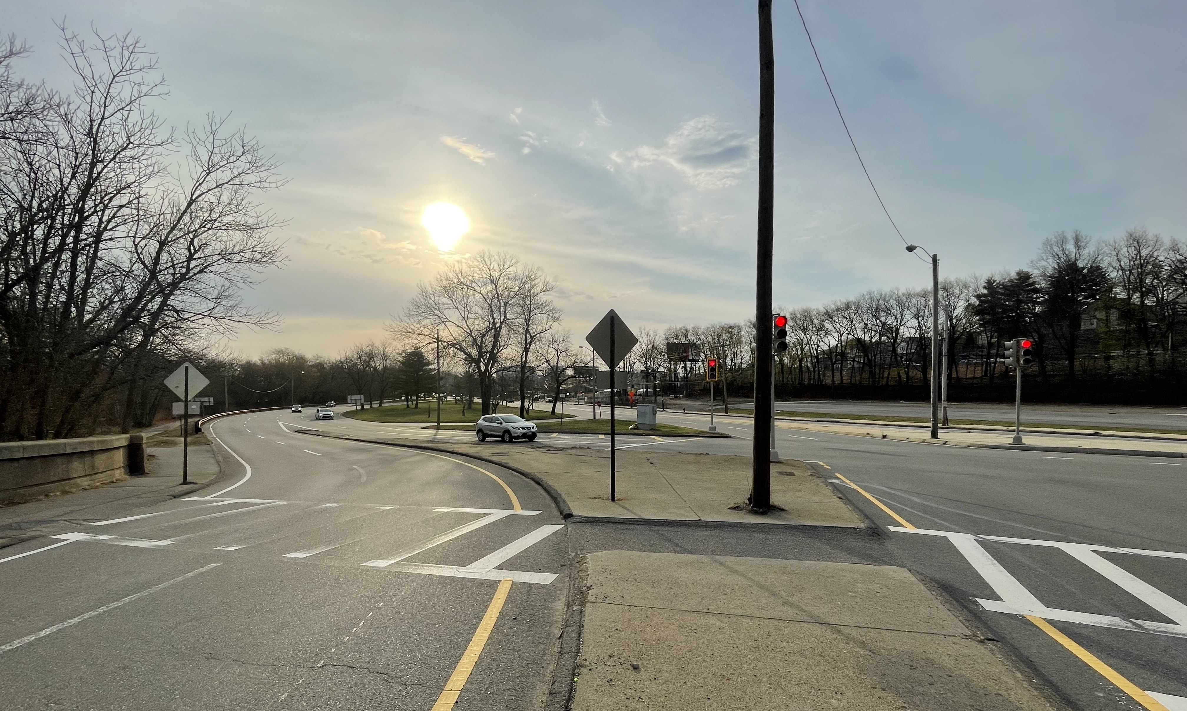 A wide intersection of several multi-lane roadways that weave around a broad traffic circle. In the foreground are two crosswalks divided by a barren concrete traffic island. At right are three red traffic lights. 