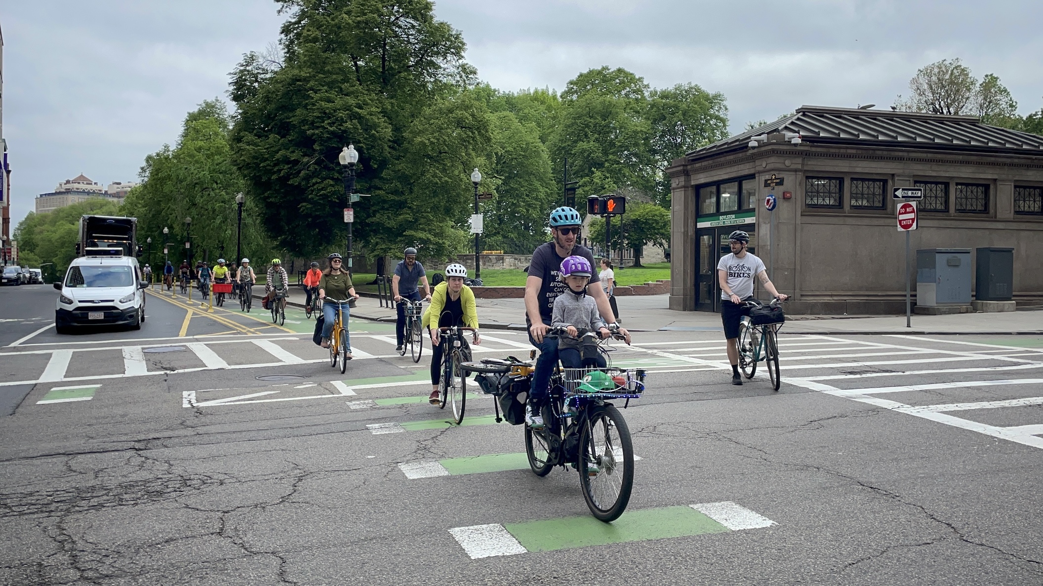 A long line of people on bikes rides along a bike lane through an intersection. In the background is a large park with a subway station entrance on the corner. A green sign on the subway station reads "BOYLSTON". In the front of the line is a man in a teal bike helmet carrying a young child in a purple helmet on a kids seat in front of him.