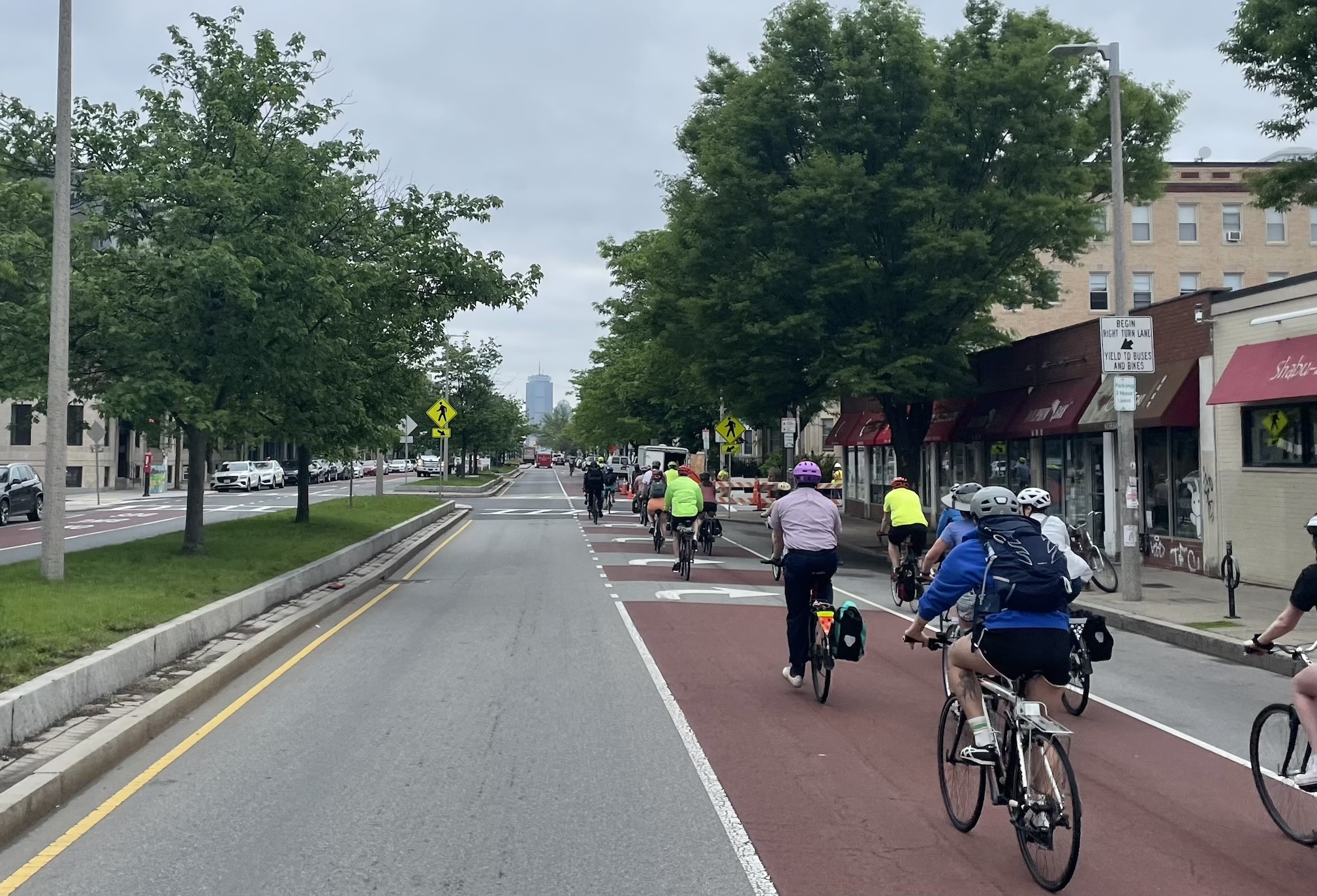 A crowd of people on bikes ride one- and two-abreast along a red-painted bus and bike lane along a multi-lane city street lined with trees. In the distance, a single skyscaper, the Prudential Center, is visible on the horizon.