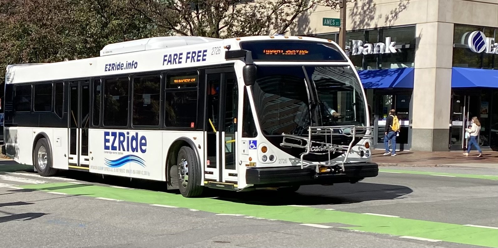 A white bus with the words "EZRide" and "FARE FREE" on its sides passes a bank and a street sign that says "Ames St" on a street with a bike lane