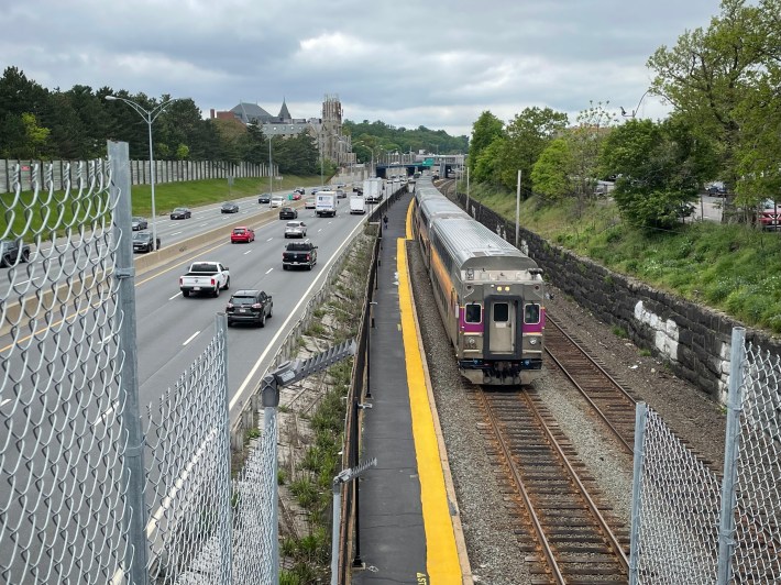 An MBTA commuter rail train, seen from a highway overpass. To the left of the train is a yellow-striped boarding platform, and to the left of the platform is a six-lane interstate highway.