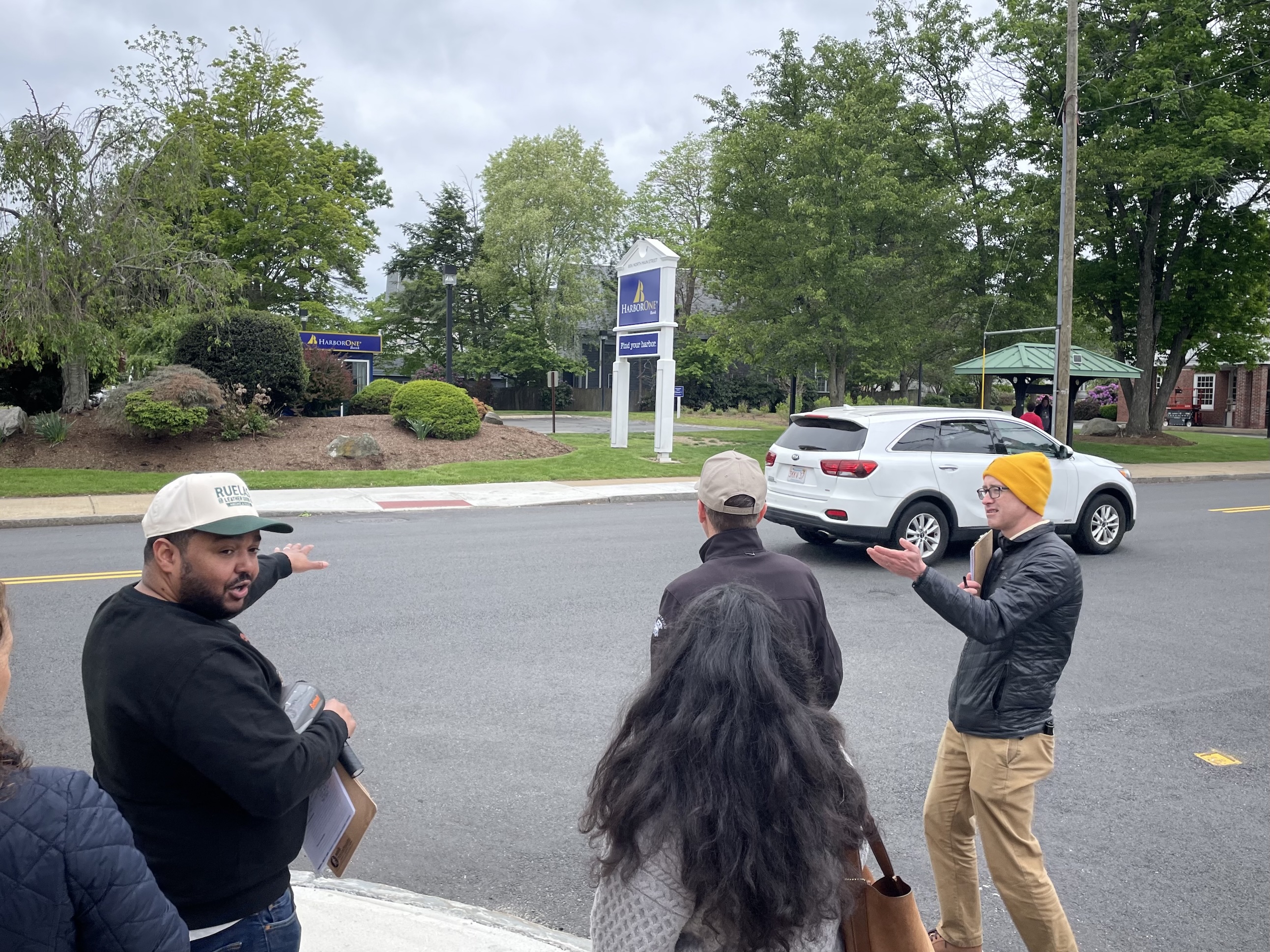 A man in a black jacket and yellow hat gestures at a city street next to three other people standing on a sidewalk.