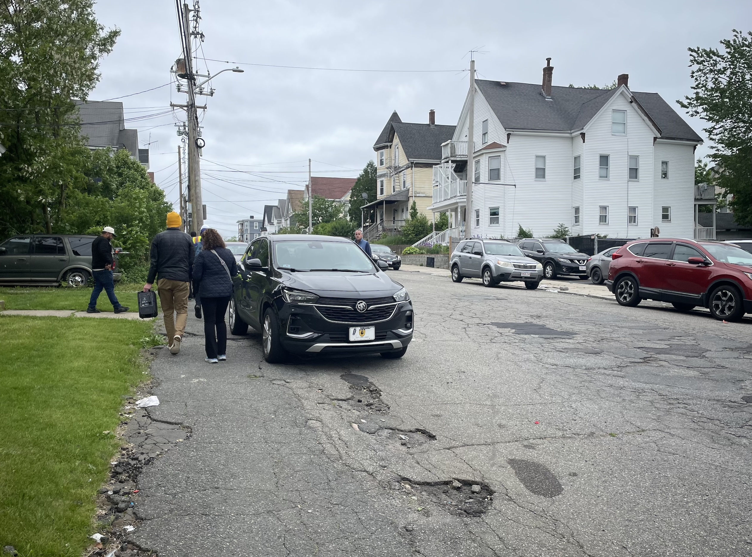 A city street with lots of potholes, lined with parked cars. Two pedestrians squeeze between a parked car and the edge of the asphalt at left.
