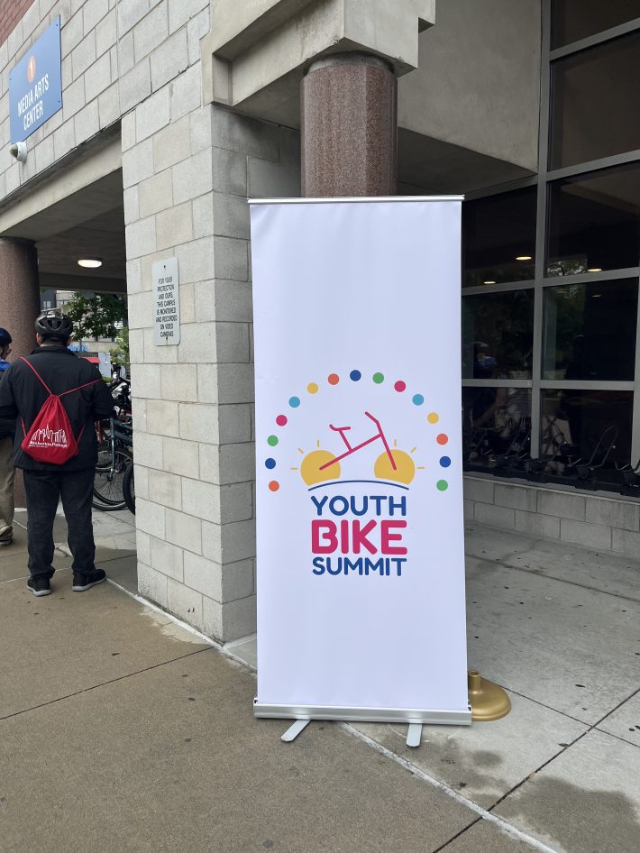 A vinyl banner for the Youth Bike Summit stands outside the Media Arts Center at Roxbury Community College. The banner features a colorful bicycle logo surrounded by a dotted semicircle.