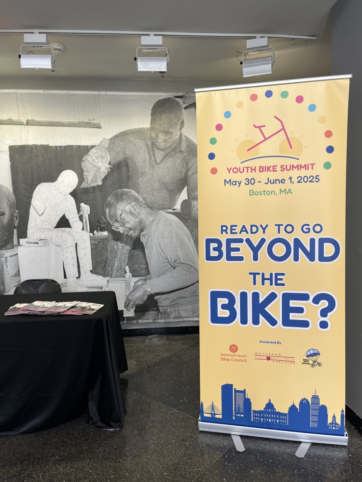 A large yellow banner at the Youth Bike Summit reads “READY TO GO BEYOND THE BIKE?” with event details for May 30–June 1, 2025, in Boston, MA. It stands next to a table with flyers and a mural in the background showing Black artists.