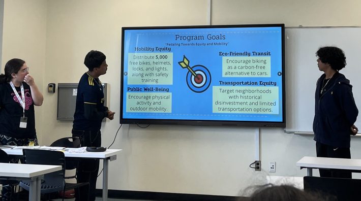 Three young people stand beside a projected slide labeled “Program Goals,” which highlights themes like Mobility Equity, Public Well-Being, Eco-Friendly Transit, and Transportation Equity.