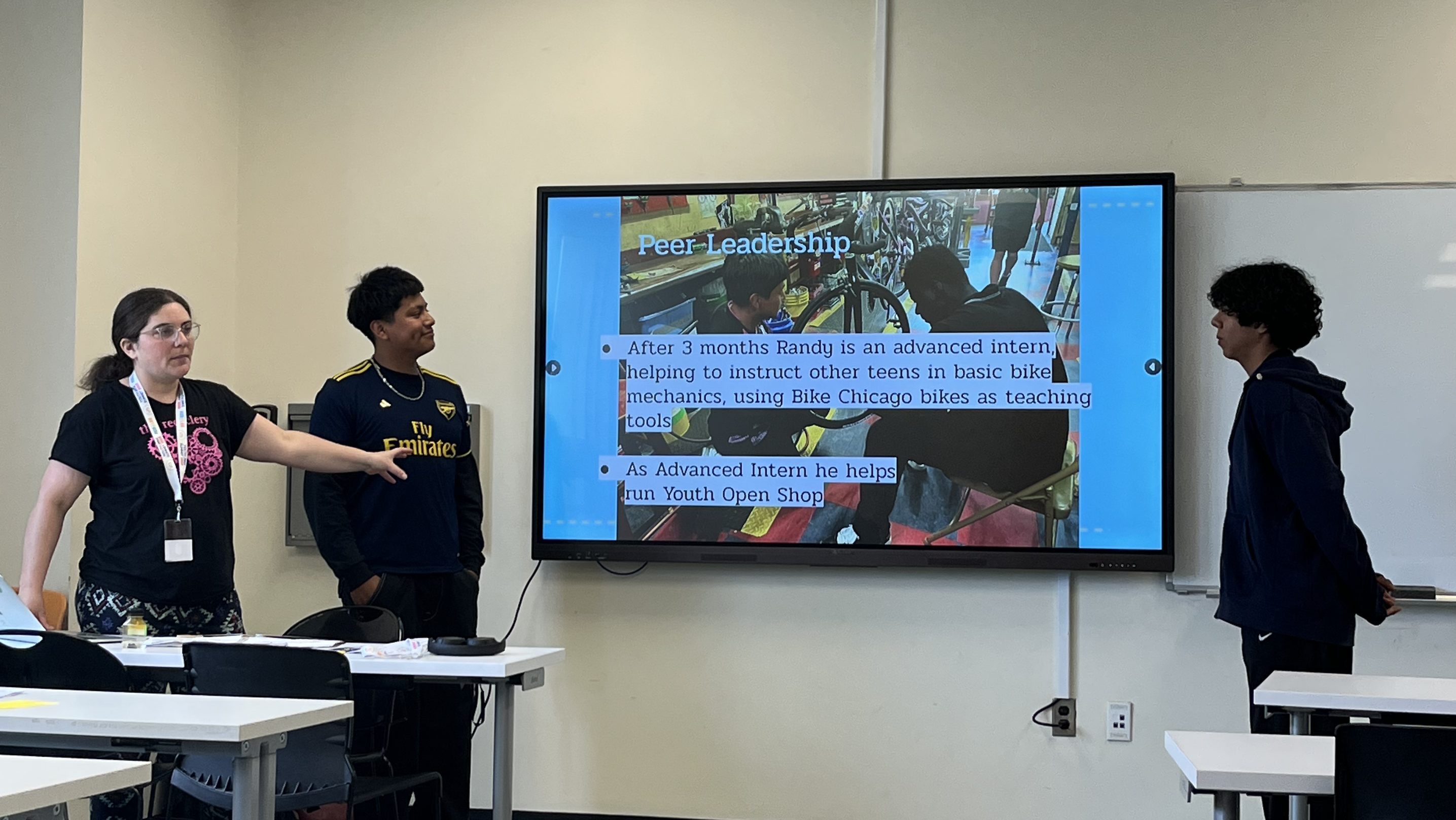 Three young people present a slide titled “Peer Leadership” about youth interns teaching bike mechanics, with the audience seated at desks.