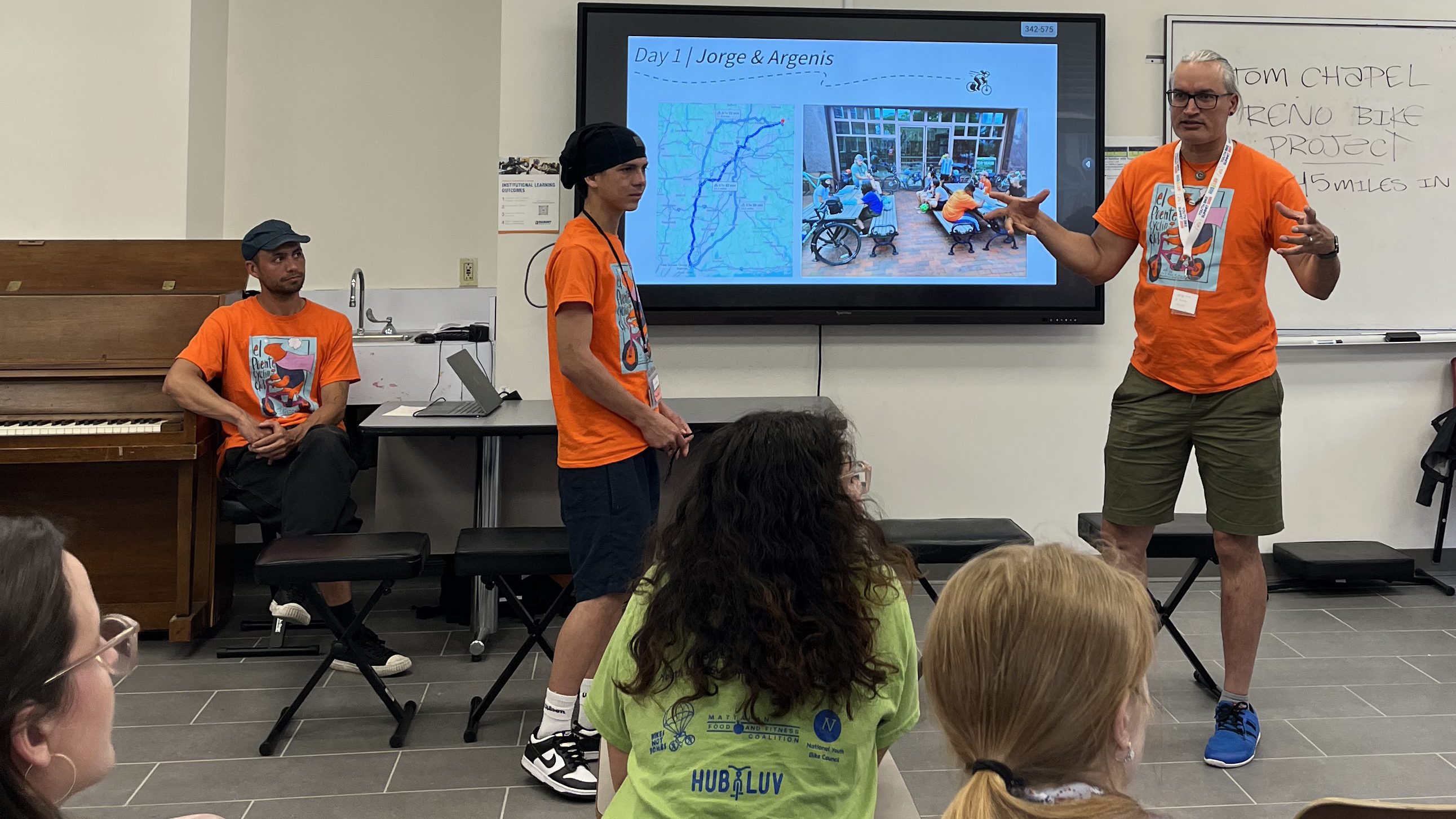Two presenters in orange shirts lead a classroom session with a slide showing a map and photo from their bike journey, titled “Day 1: Jorge & Argenis,” while attendees listen.