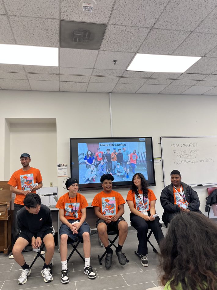 A panel of five youth wearing bright orange El Puente shirts sit in front of a classroom. A slide behind them says “Thanks for coming!” with a group photo.