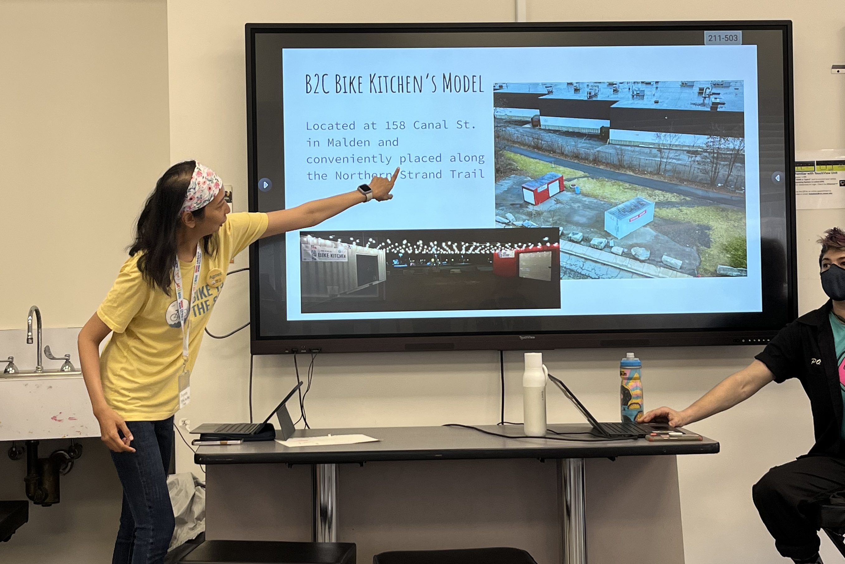 A presenter points to a slide about the B2C Bike Kitchen model in Malden, MA, highlighting its location on the Northern Strand Trail.