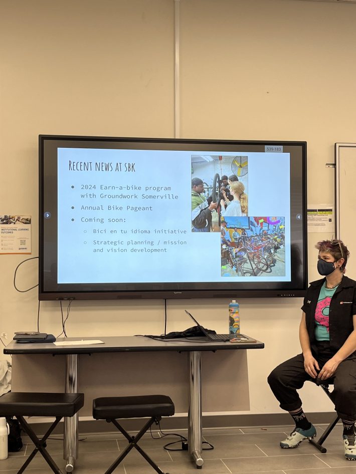 A short-haired woman in a mask sits next to a projector screen with the words "recent news at SBK: 2024 earn-a-bike program with Groundwork Somerville; Annual Bike Pageant; Coming soon: Bici en tu idioma initiative and Strategic planning"