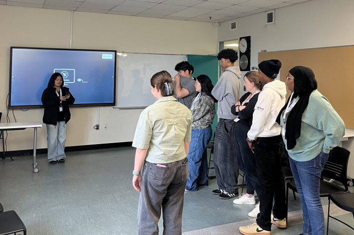 A group of youth from the ECO Youth session stand in a semicircle at the front of a classroom, smiling and talking after presenting.