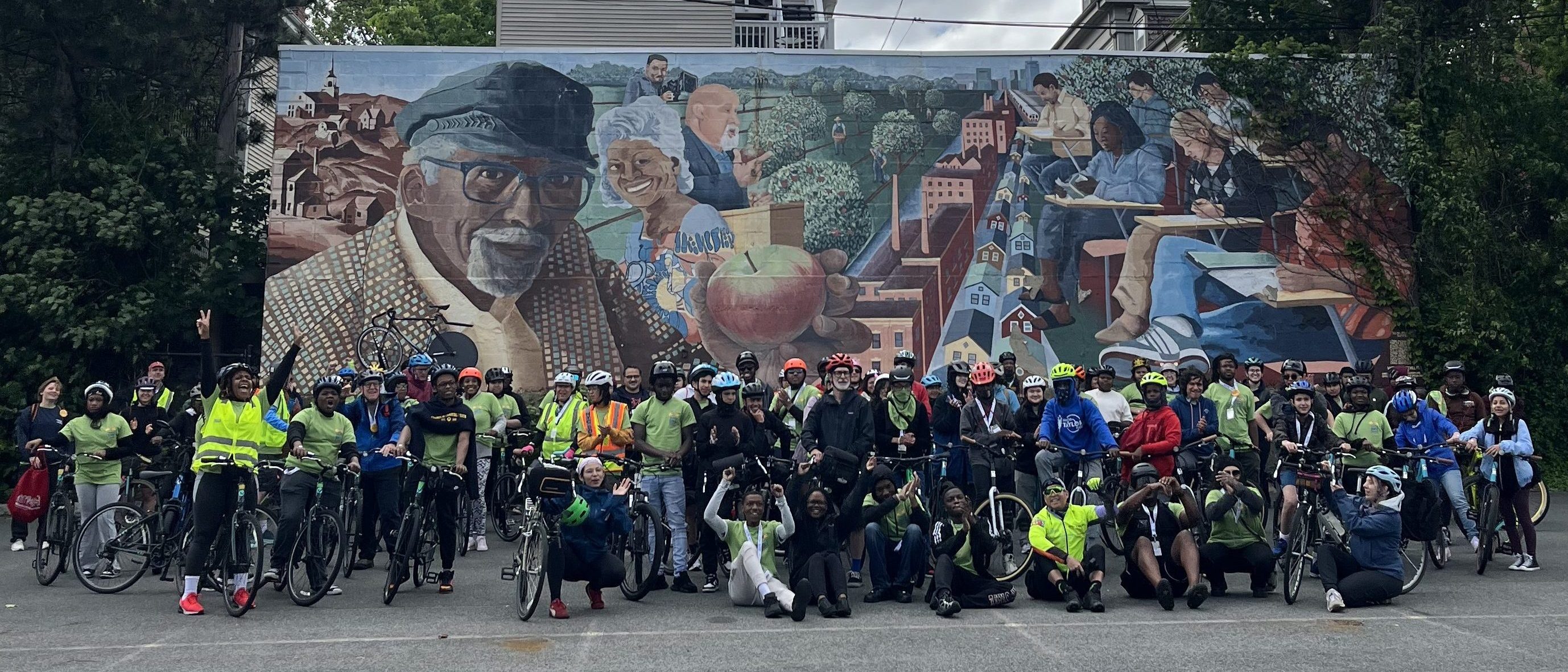 A large group of cyclists, many wearing helmets and bright clothing, pose in front of a vibrant community mural that features portraits of Black elders, children, and neighborhood scenes, set in a parking lot surrounded by trees and houses under a cloudy sky.