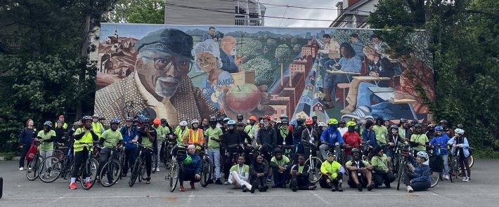 A large group of cyclists, many wearing helmets and bright clothing, pose in front of a vibrant community mural that features portraits of Black elders, children, and neighborhood scenes, set in a parking lot surrounded by trees and houses under a cloudy sky.