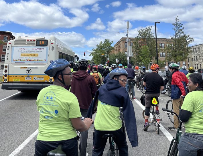 A crowd of cyclists wait at a traffic light behind an MBTA Route 16 bus on a major Boston street, wearing colorful helmets and event shirts.