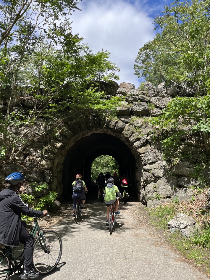 Bicyclists ride through a rocky tunnel entrance surrounded by greenery on a paved path.