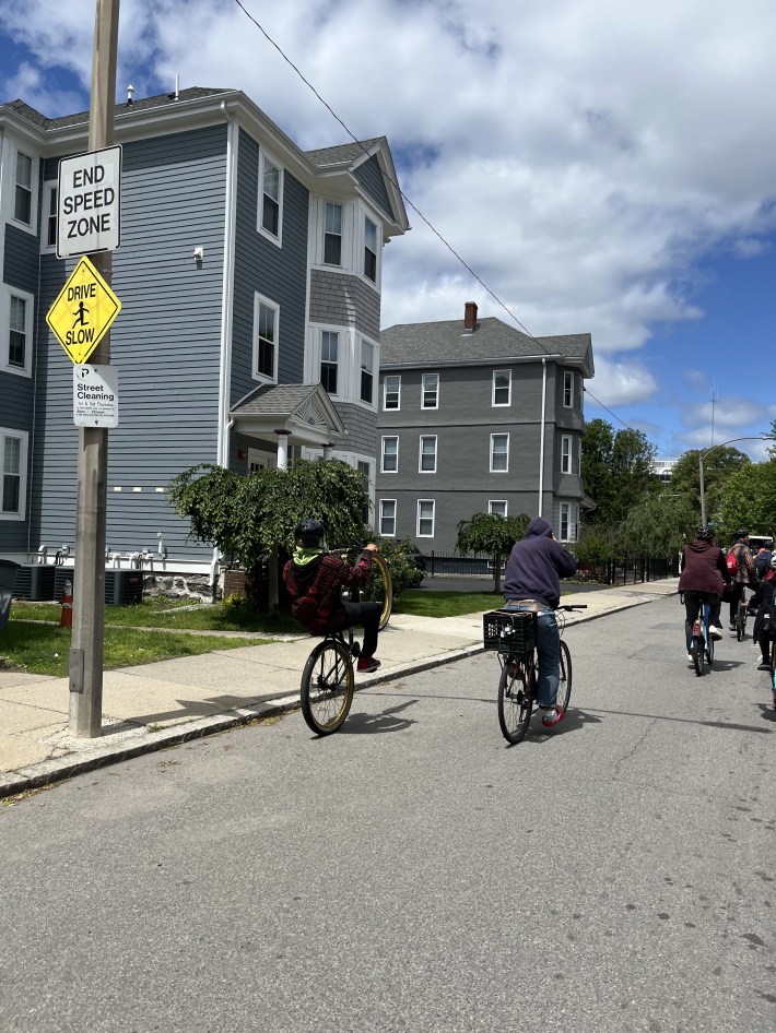 Riders cycle past residential houses on a neighborhood street marked with a “Reduced Speed Zone” sign.