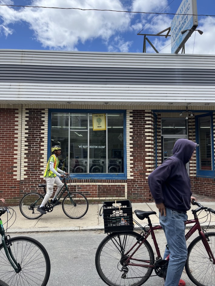 Youth and adults on bikes ride past a brick laundromat in Boston, including one cyclist in a reflective vest and helmet, and another with a crate mounted on the back of their bike.