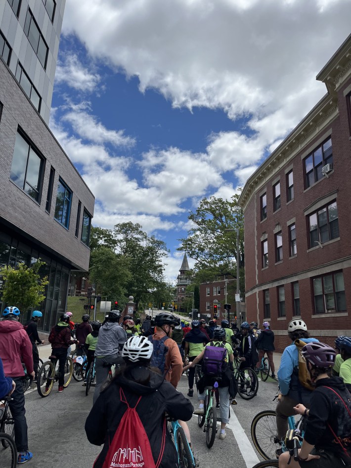 A large group of cyclists, many wearing helmets and backpacks, ride through a city street with brick buildings under a partly cloudy sky.