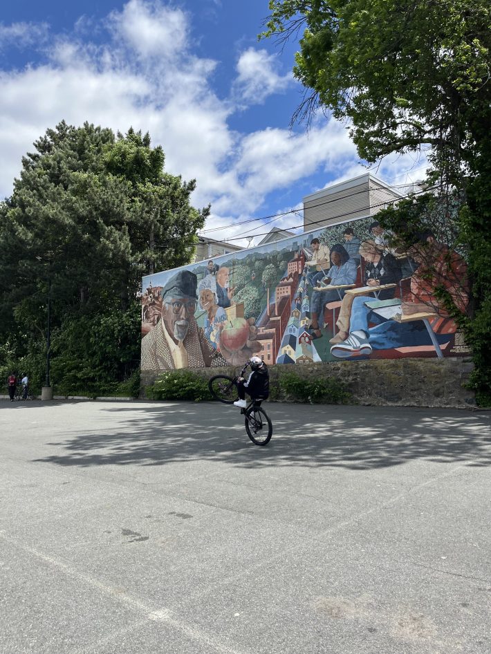 A cyclist performs a wheelie in front of the vibrant “History of Fort Hill, Pt.2” mural, which features scenes of Roxbury’s community history.