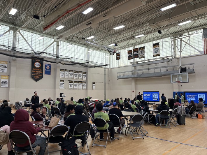 Participants wearing green Youth Bike Summit shirts gather in a gymnasium for a group session, sitting at round tables in front of presentation screens.