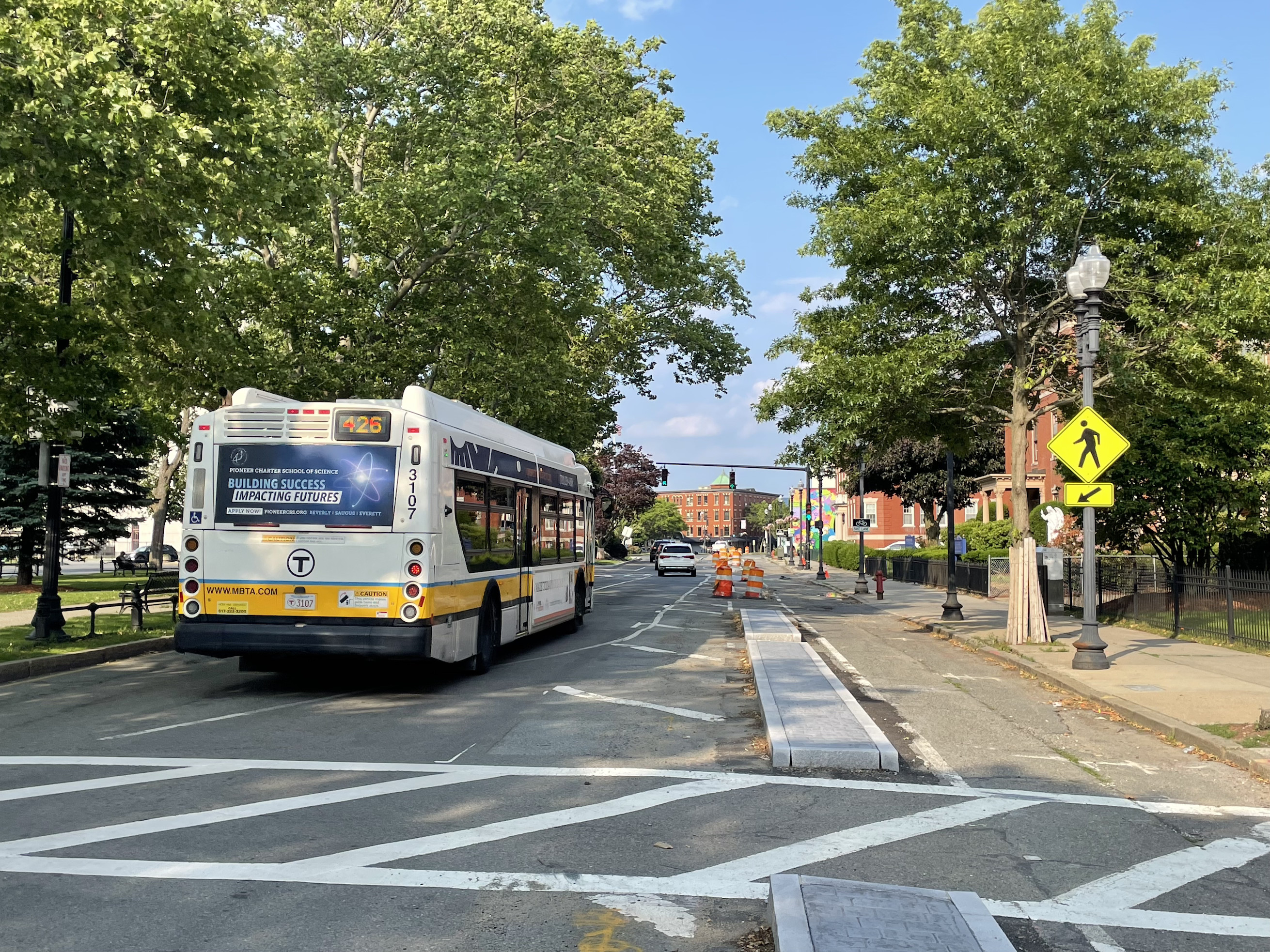 A city street lined with large mature deciduous trees. In the foreground are an MBTA bus (left) and a crosswalk sign (right). A curbed median divides the street with two lanes for cars on the left and a bike path and sidewalk to the right.