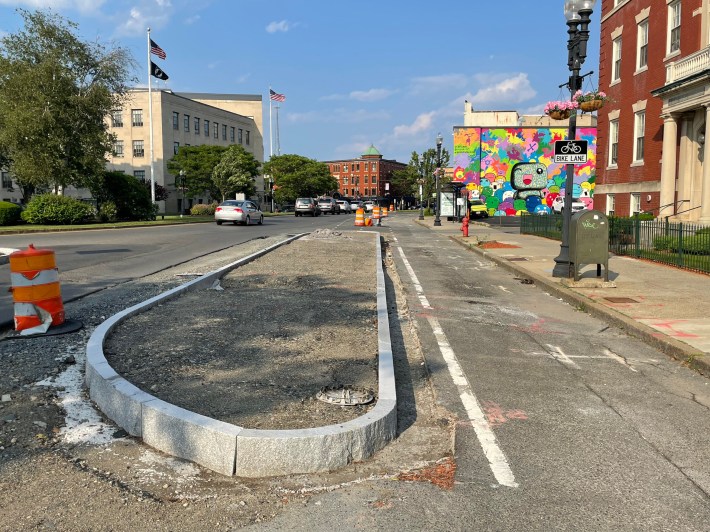 A city street lined with older masonry multi-story buildings, including one with a colorful wall mural. In the foreground is a large curbed median island that divides the street with two lanes for cars on the left and a bike path and sidewalk to the right.