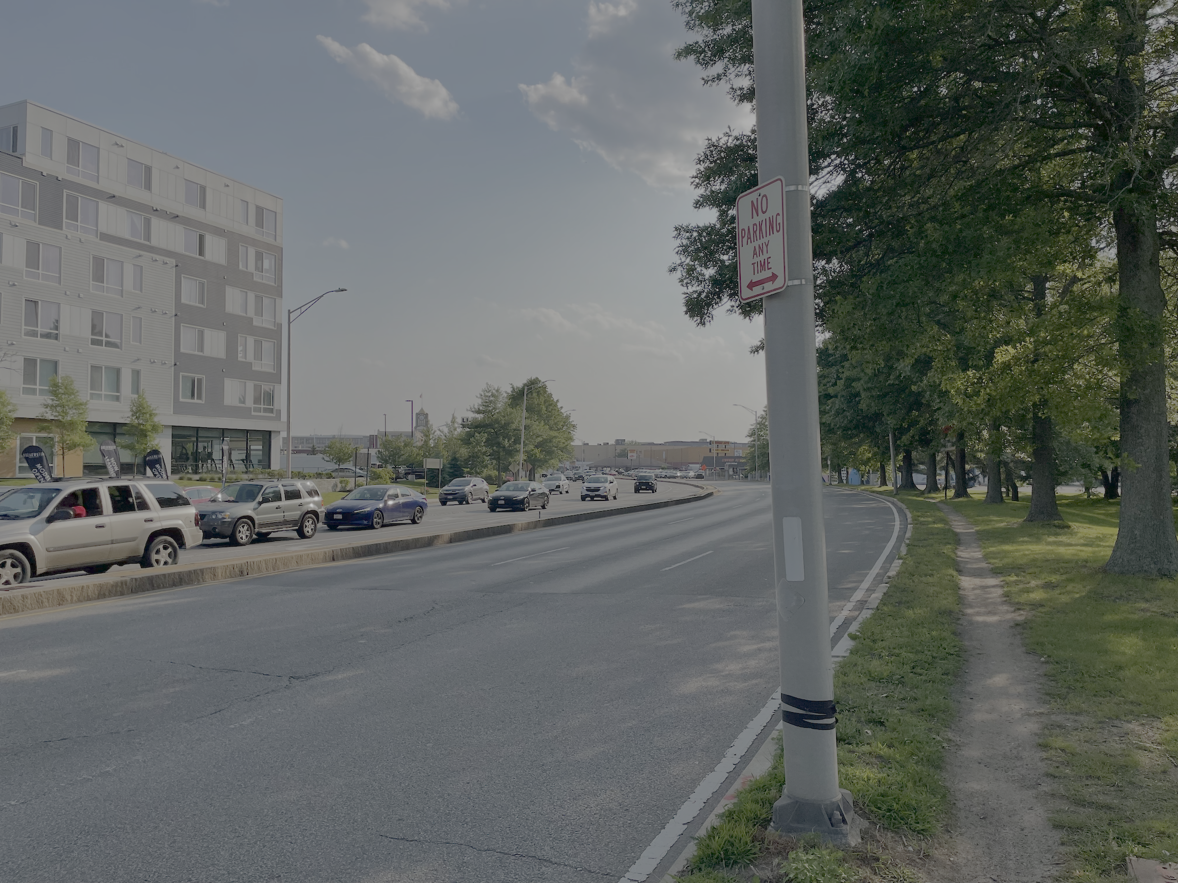 A well-worn dirt path runs alongside an allee of trees next to a large 6-lane-wide divided highway. On the left edge of the photo, on the opposite side of the roadway, is a 6-story apartment building. A metal street lamp pole in the foreground bears a "no parking any time" sign.