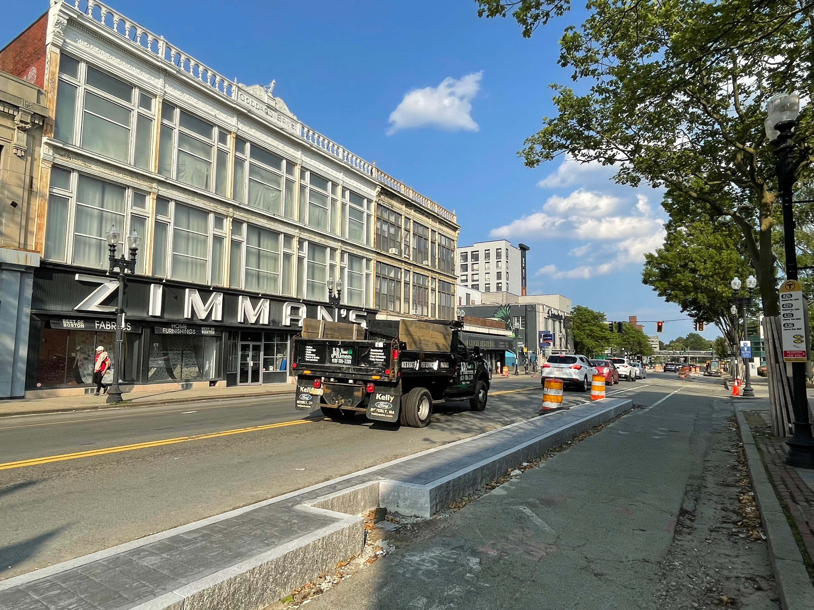 A city street with a n ornate three-story building on the opposite side with a large sign reading "ZIMMAN'S" above its ground-floor storefront. In the foreground a curbed median divides the street with two lanes for cars on the left and a bike path and sidewalk to the right.