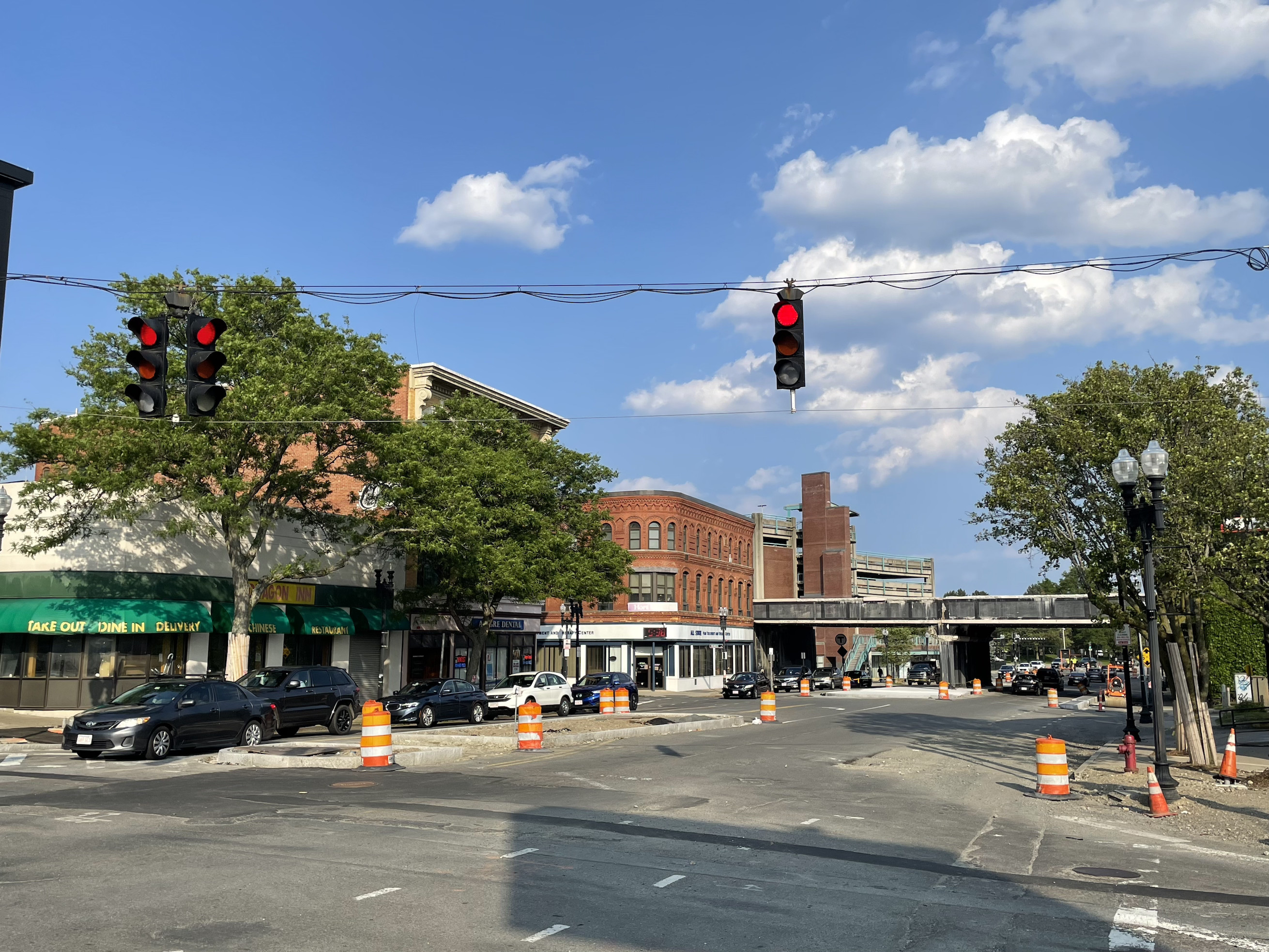 A city intersection under construction with several orange cones and construction barrels. On the opposite side of the intersection are several multi-story brick buildings and in the distance is a railroad bridge over the street.