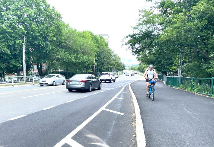 A man riding a bike on a widen path next to a wide multi-lane roadway lined with leafy trees